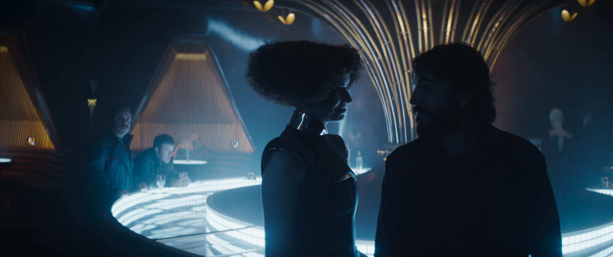 A Man talks to a Woman at a dimly lit bar. In the background a man sits, while his partner stands looking at them.