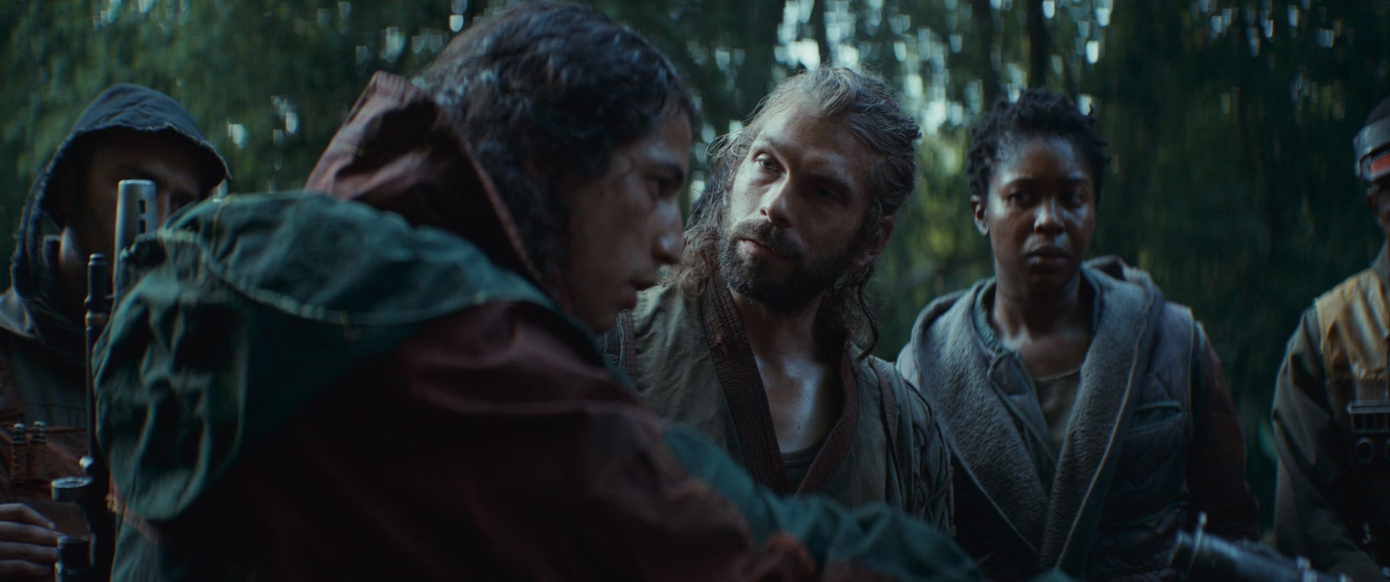 A group of bedraggled people in irregular military gear stand near each other tensely in the middle of a jungle.