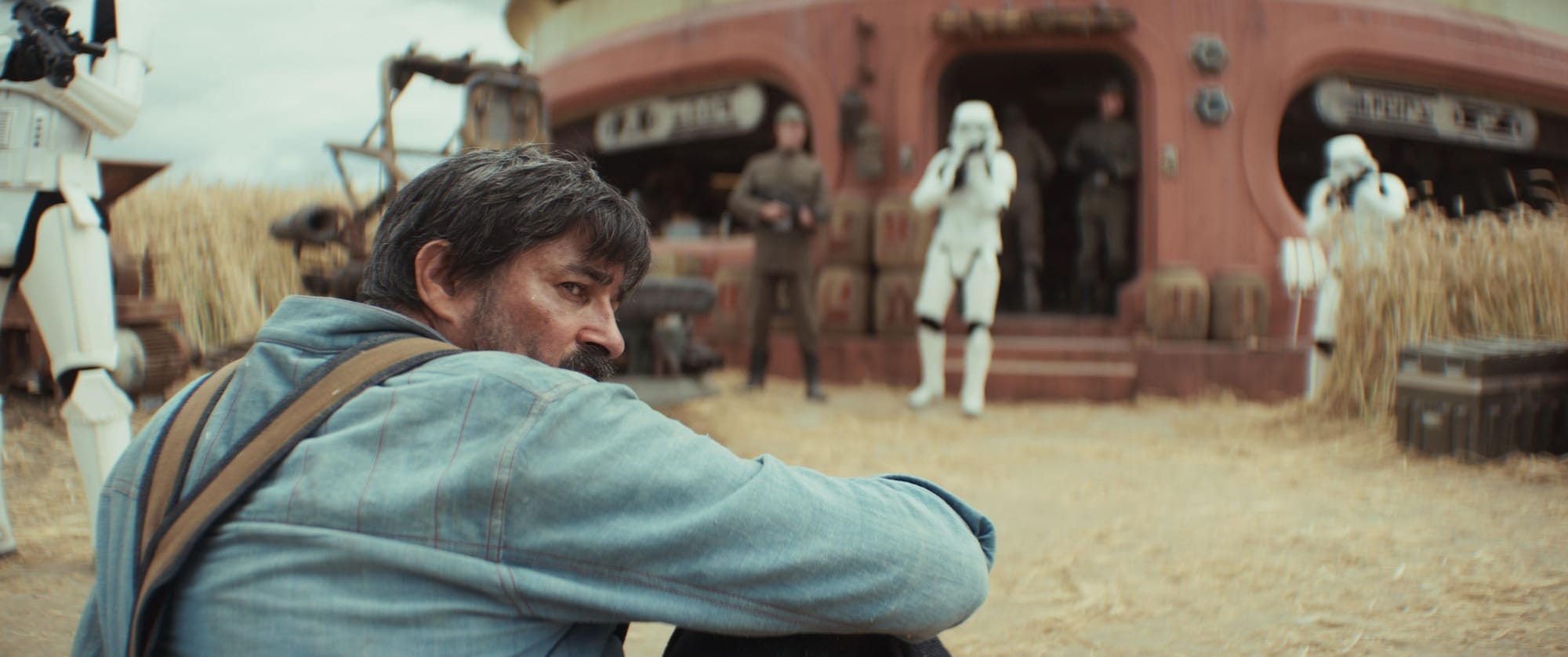 A man in a blue work shirt and denim pants sits on the ground in front a general store surrounded by stormtroopers.