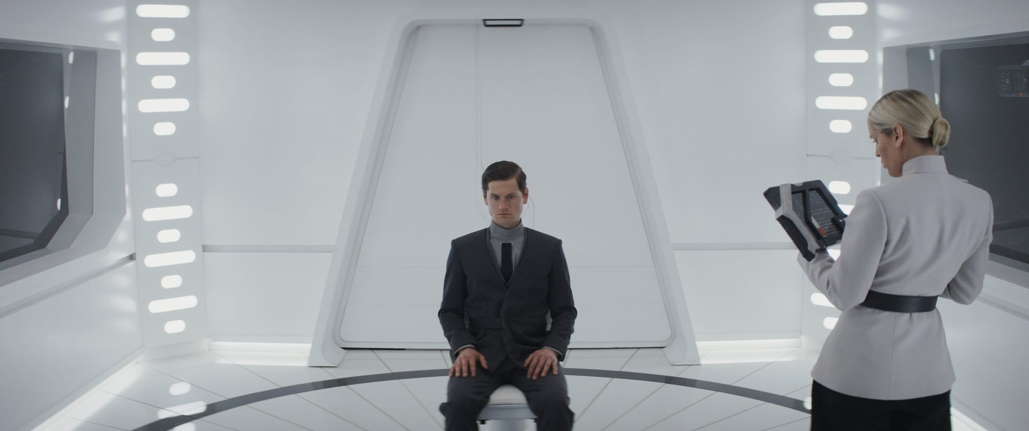 A man in a dark gray suit sits in a chair in the center of a white room. A woman in a white uniform stands near him looking at a data tablet.