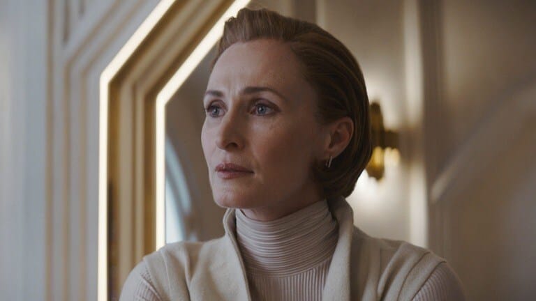A middle-aged woman with red hair in a white outfit looks off into the distance with a concerned look.