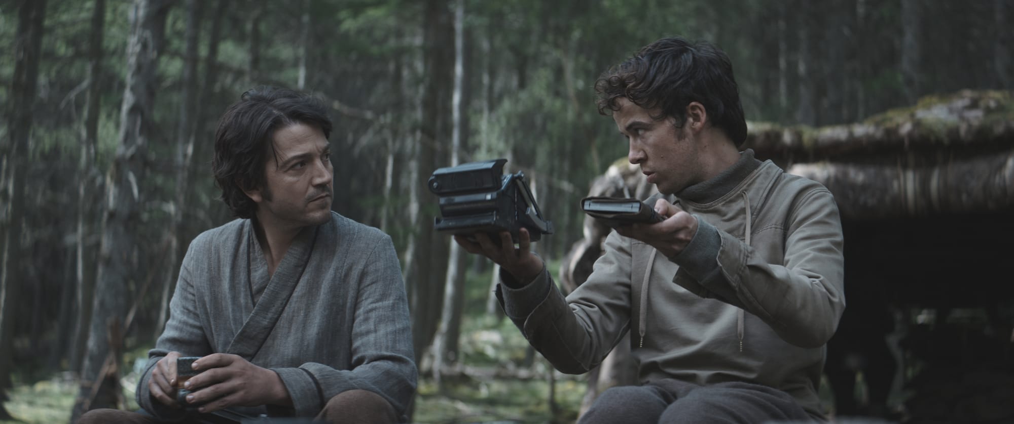 A young man in a gray tunic holds a writing pad and a nav device. He is sitting and talking to Cassian Andor.