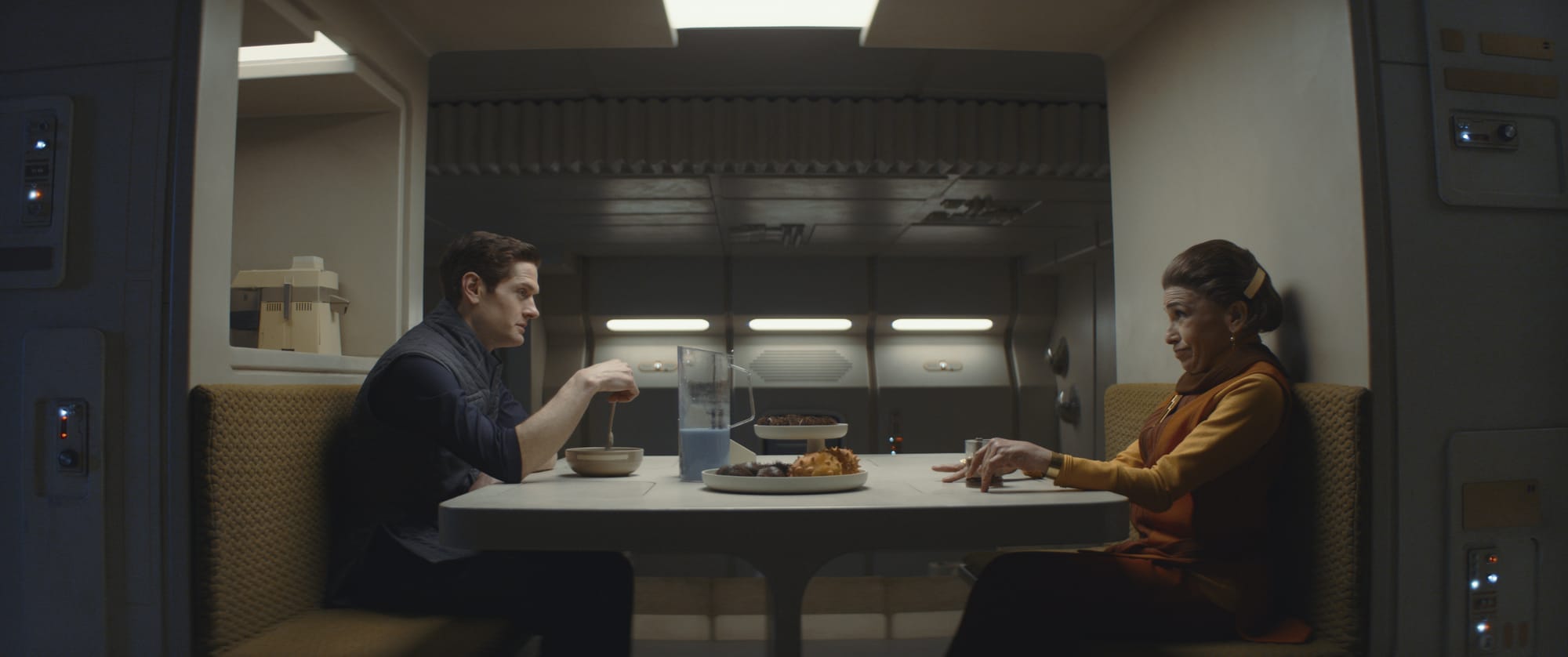 A man sits across from his mother at a table in her kitchen. He looks annoyed, and she looks like he should visit more often.