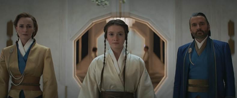 A young girl with braided hair in a white robe stands between her mother in a yellow coat and blue dress, and her father in a blue coat and robe. 