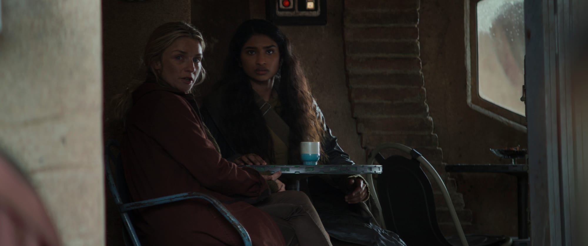 A woman in a red coat and a woman in a dark brown coat sit at a table in a cafe. They are looking at someone off screen to the front in a way not to arouse suspicion.