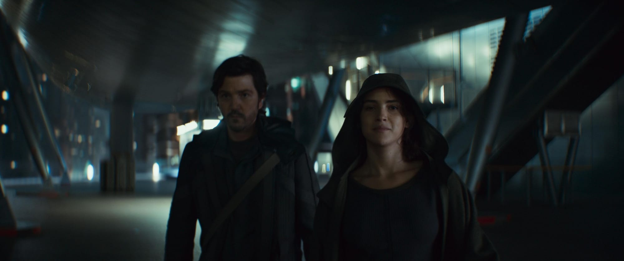 A woman in a hooded coat walks away from a futuristic looking building with a man in a dark jacket