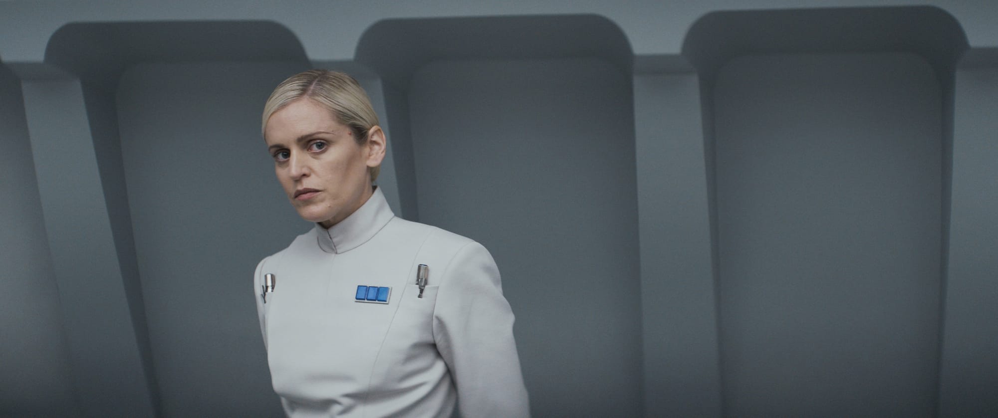 A woman in a gray uniform stands in a sterile gray room and looks skeptically at something off screen.