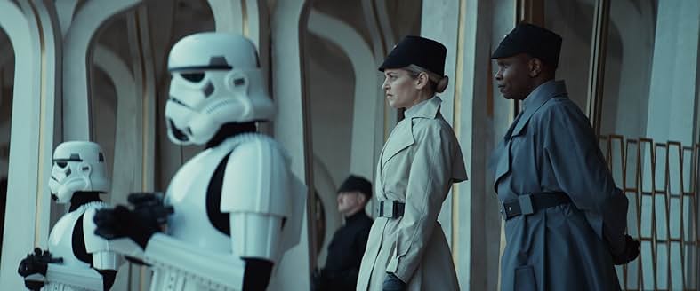 A woman in an off white coat and dark Imperial cap stands behind a pair of stormtroopers as her gray coated lieutenant stands next to her.