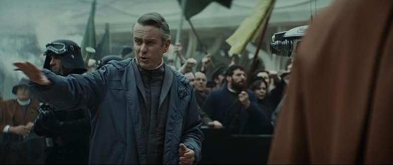 A man in a blue coat stands in front of a hovering camera droid reporting on the protest happening around him.