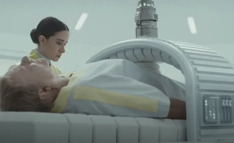 A man is unconscious on a white bed. A machine is around his torso keeping him alive. A young woman is taking it off of him.