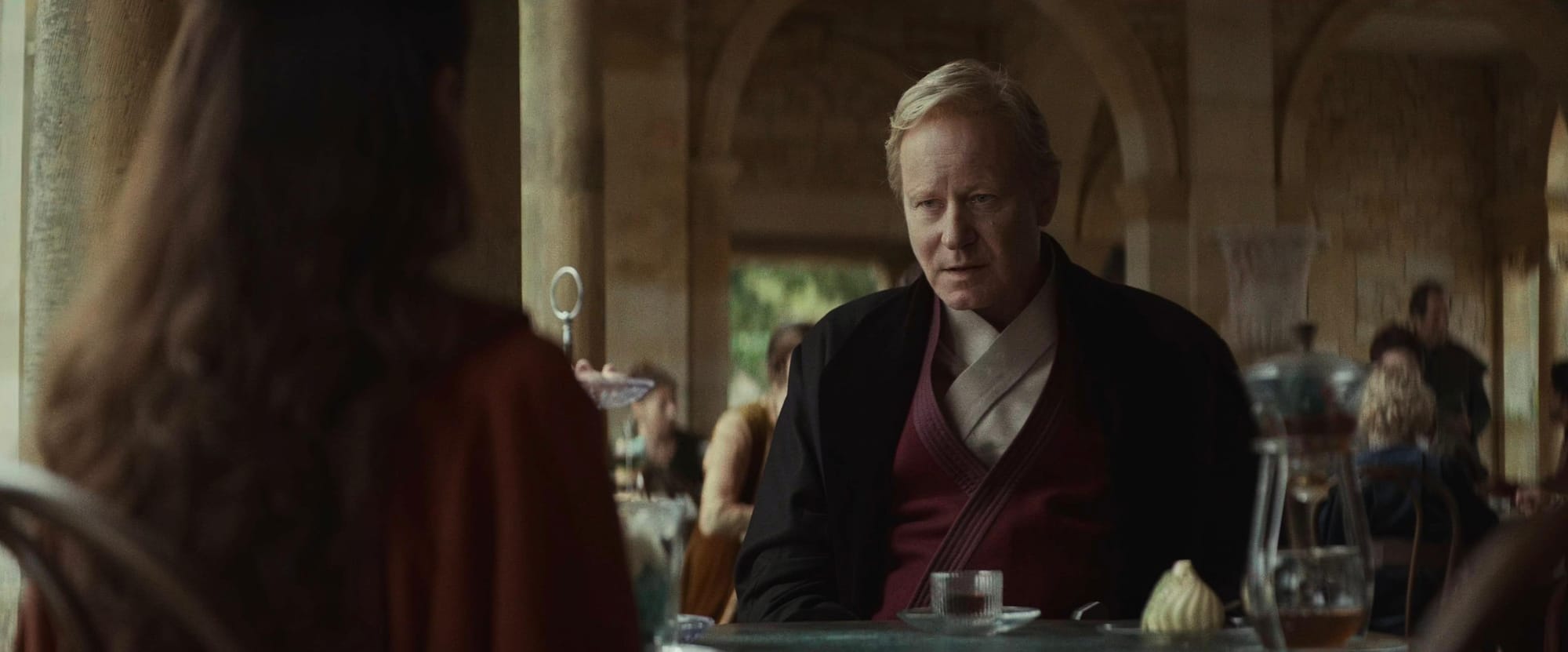 A middle-aged man with blond hair sits in an openair cafe. He wears a black coat over a brown robe and talks to a young girl.