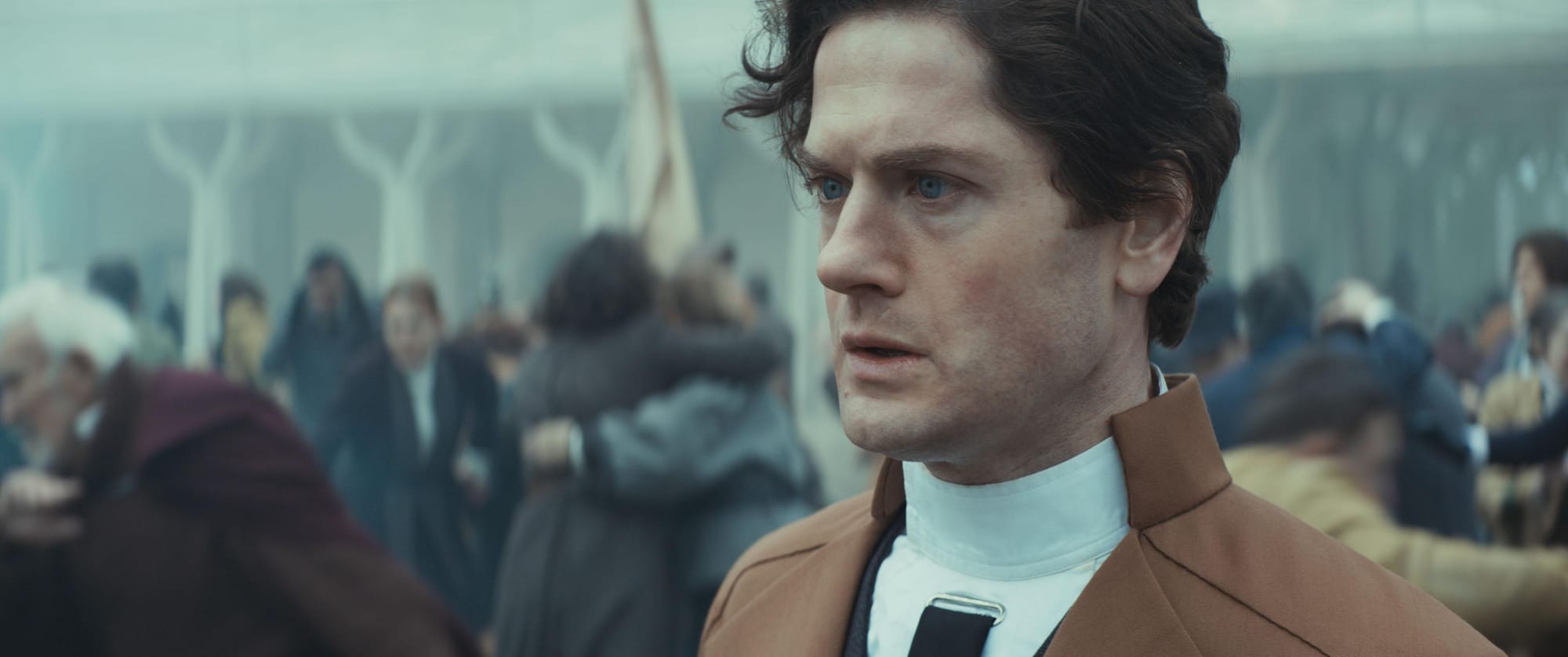 A man in a black suit and brown overcoat stands with a stunned look as people are massacred all around him.