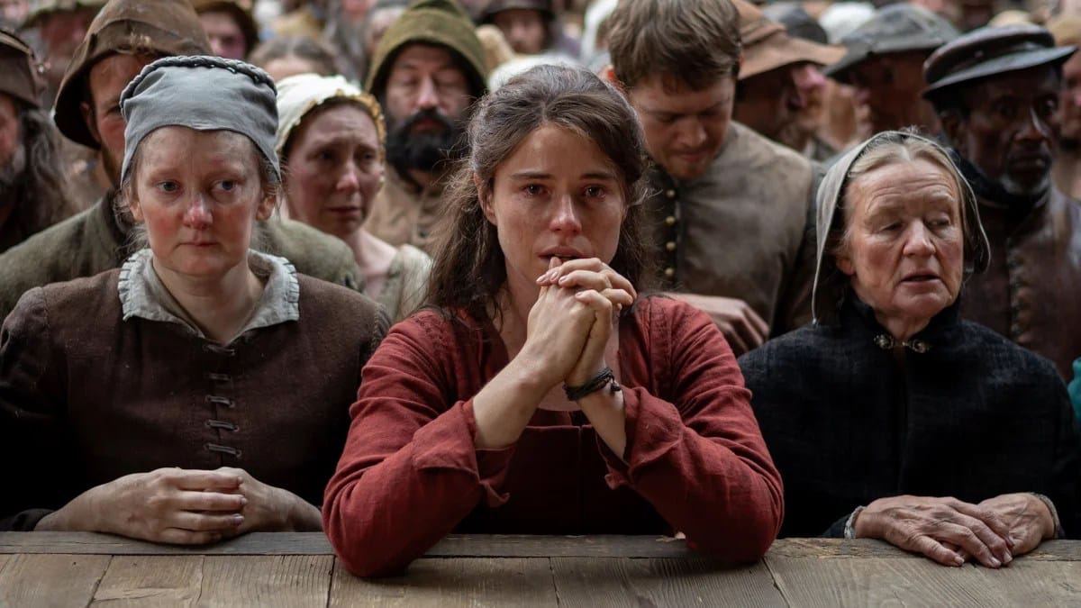 A woman with long brown hair in a red dress clasps her hands together as if at prayer. She stares intently at the play.