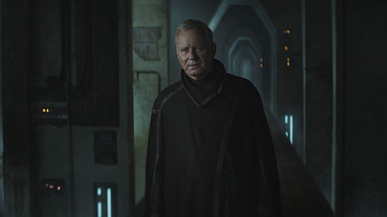 An older man in a dark cloak stands atop a dimly lit narrow walkway without any safety rails.