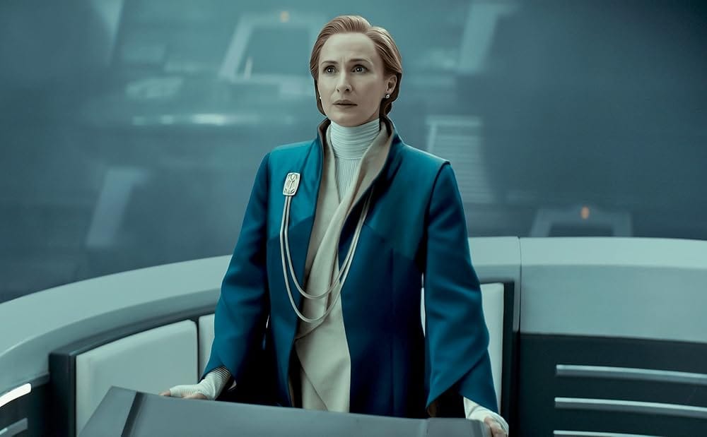 A middle aged woman with red hair stands at a podium in the middle of a Senate chamber and gives a speech. She wears a blue coat over a tan robe.