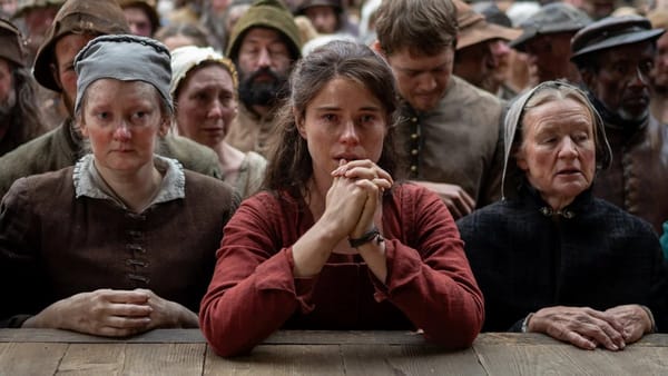 A woman with long brown hair in a red dress clasps her hands together as if at prayer. She stares intently at the play.