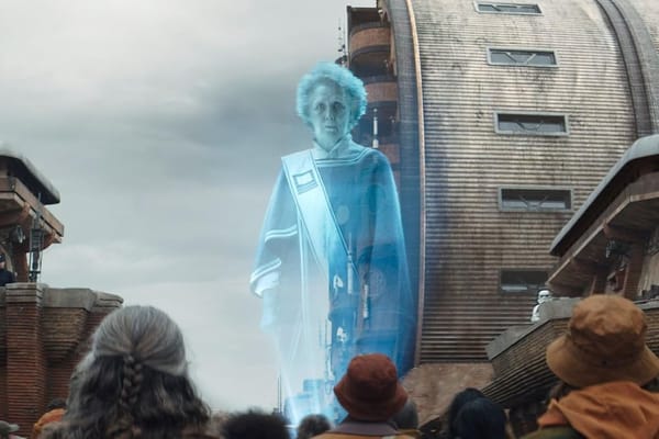 A holographic projection of an elderly woman towers over a group of people in red and orange clothing in an industrial looking city.