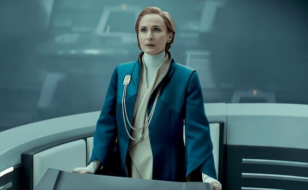 A middle aged woman with red hair stands at a podium in the middle of a Senate chamber and gives a speech. She wears a blue coat over a tan robe.