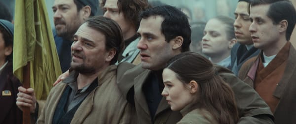 A large group of people dressed in brown and tan clothes stand closely packed in a crowd of protestors and sing.